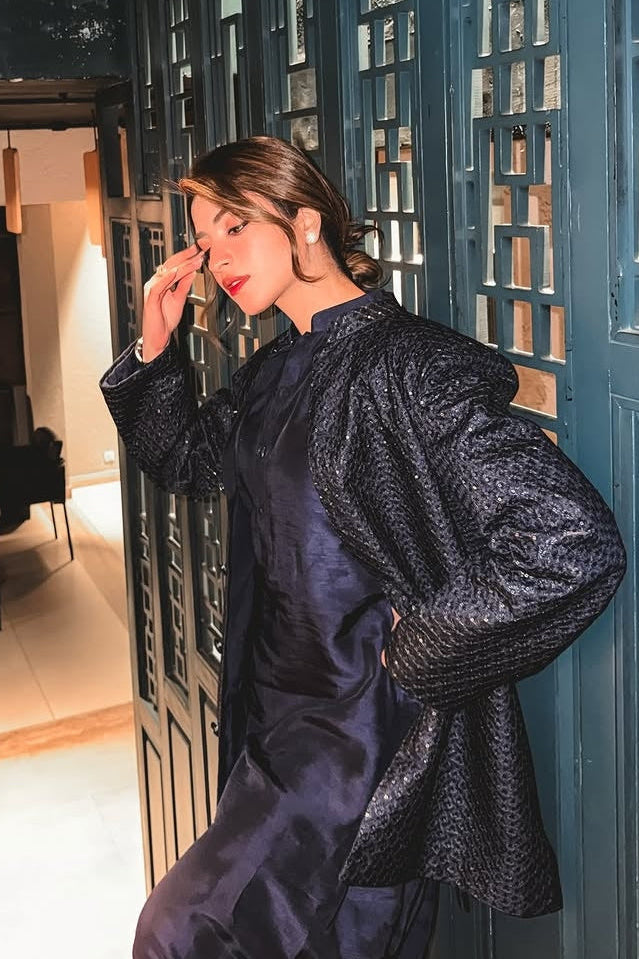 Woman in a dark blue textured coat standing in a modern interior setting with geometric patterns.
