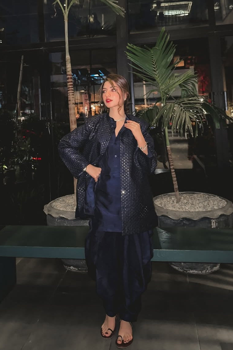 Woman in a dark blue traditional outfit standing in an indoor setting with plants and a bench.