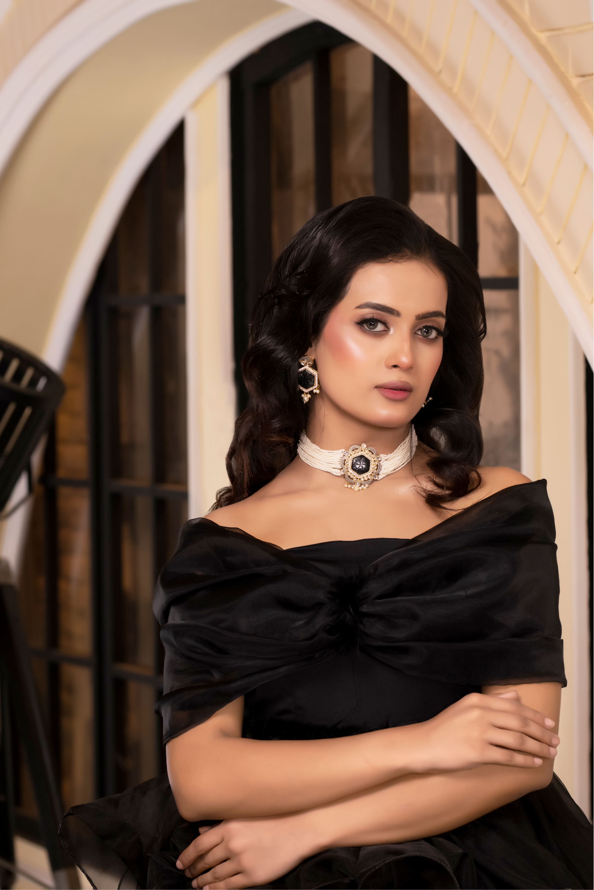 Woman in a black off-shoulder dress standing in an indoor setting with a light-colored wall and window.