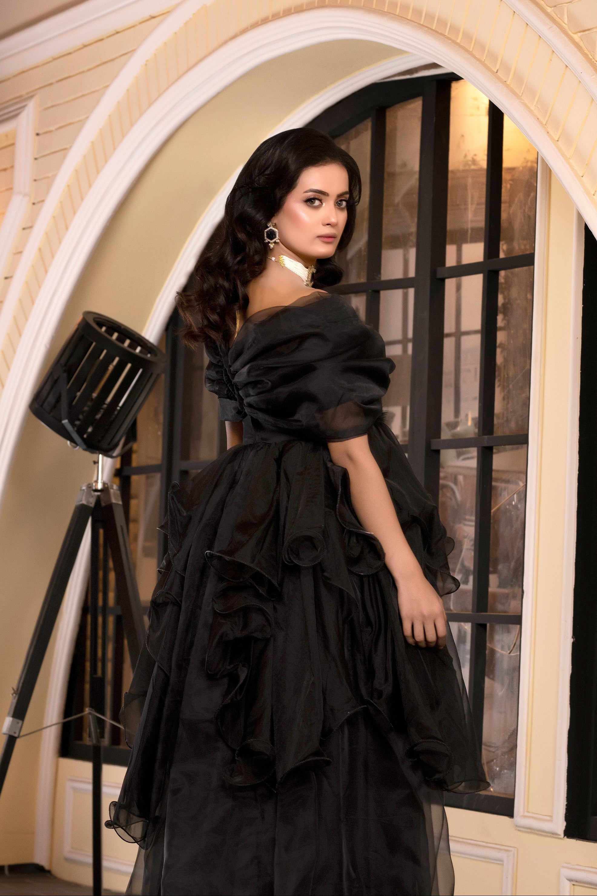 Woman in a black dress standing in an elegant interior setting with arches and lamps.