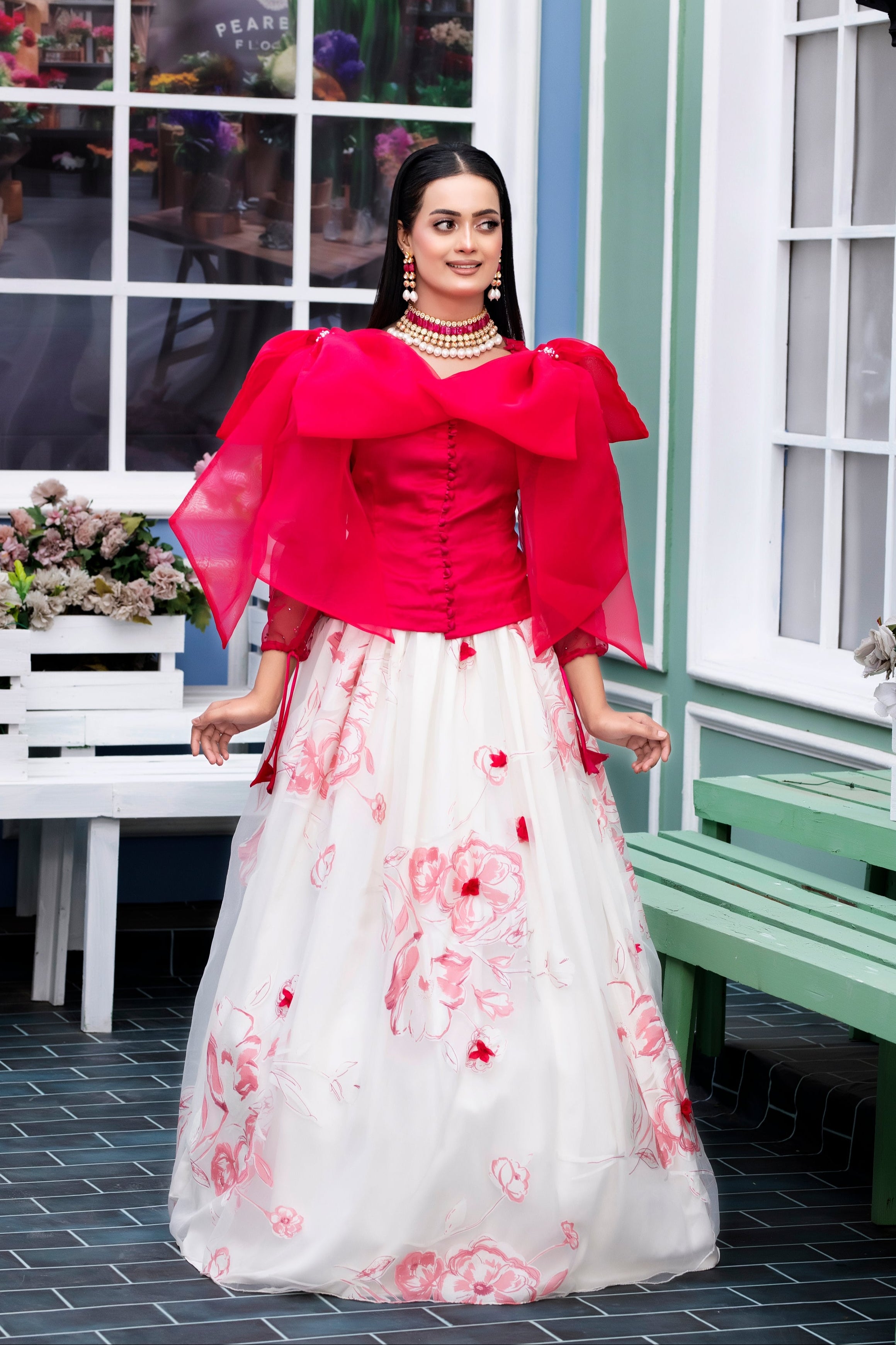 Woman in a red blouse and white floral skirt standing in front of a window with flowers.