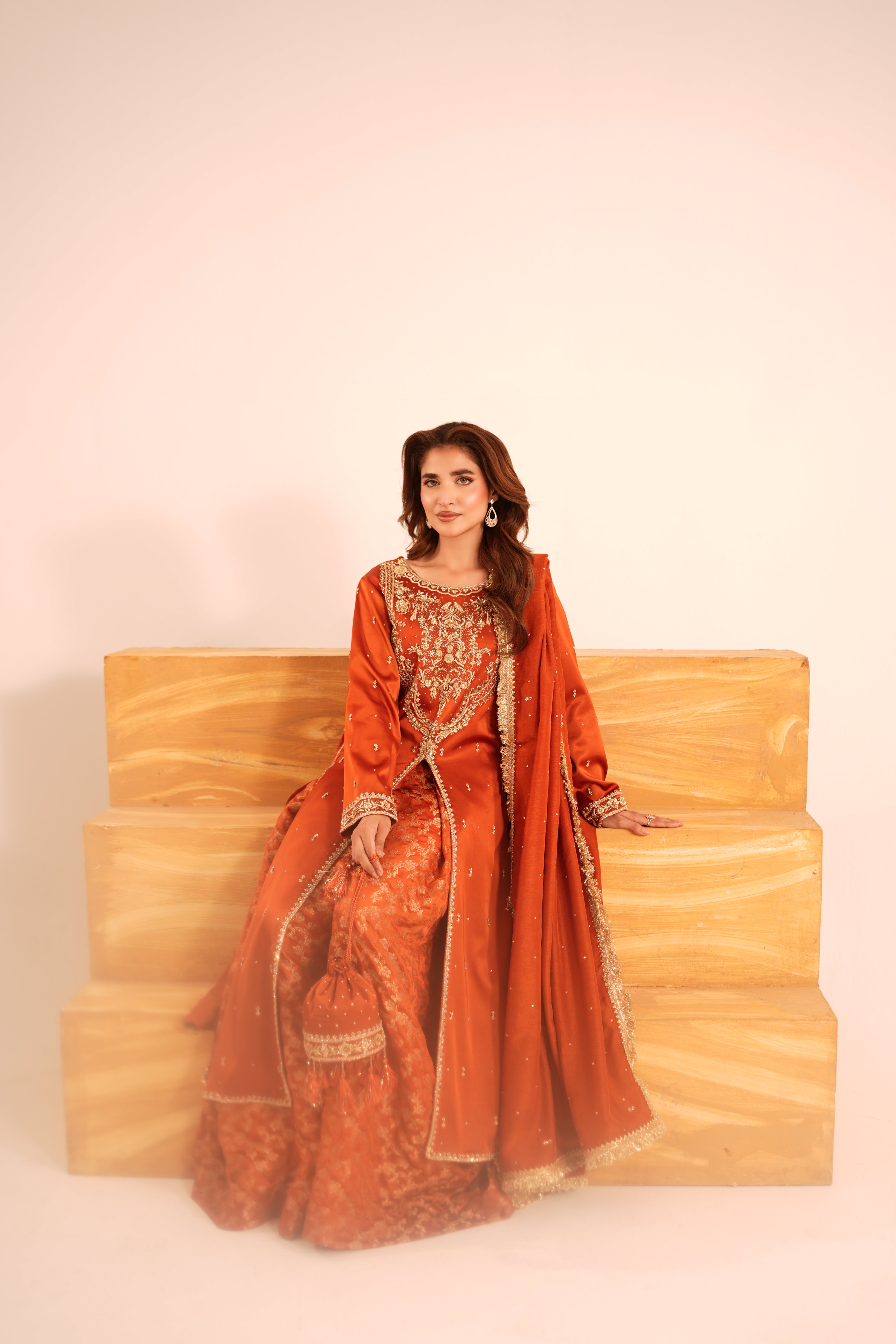 A woman wearing a traditional orange and gold embroidered dress sits on a light wood staircase against a plain off-white background.