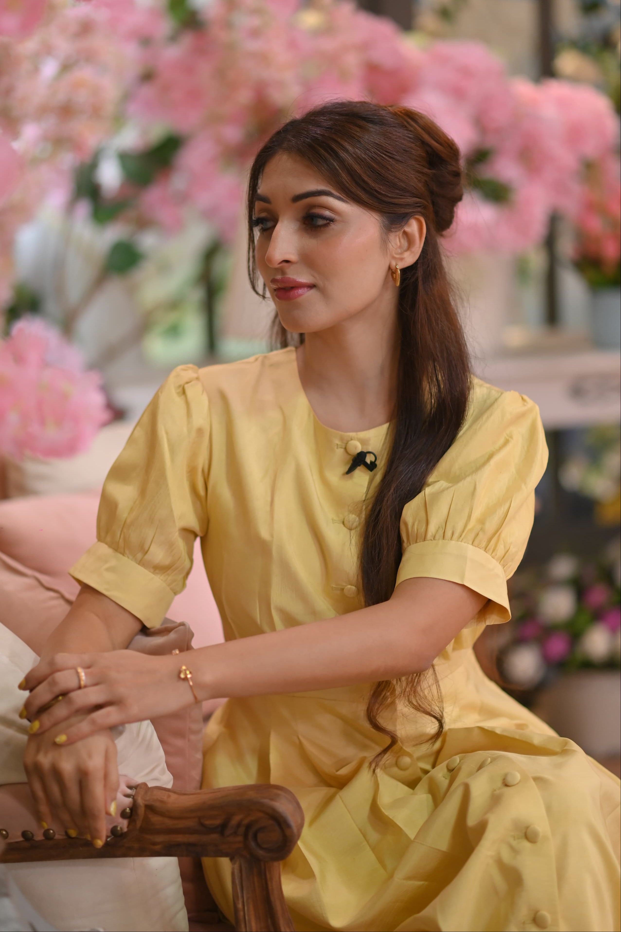 Woman in a yellow dress sitting in a decorated indoor setting with pink flowers.