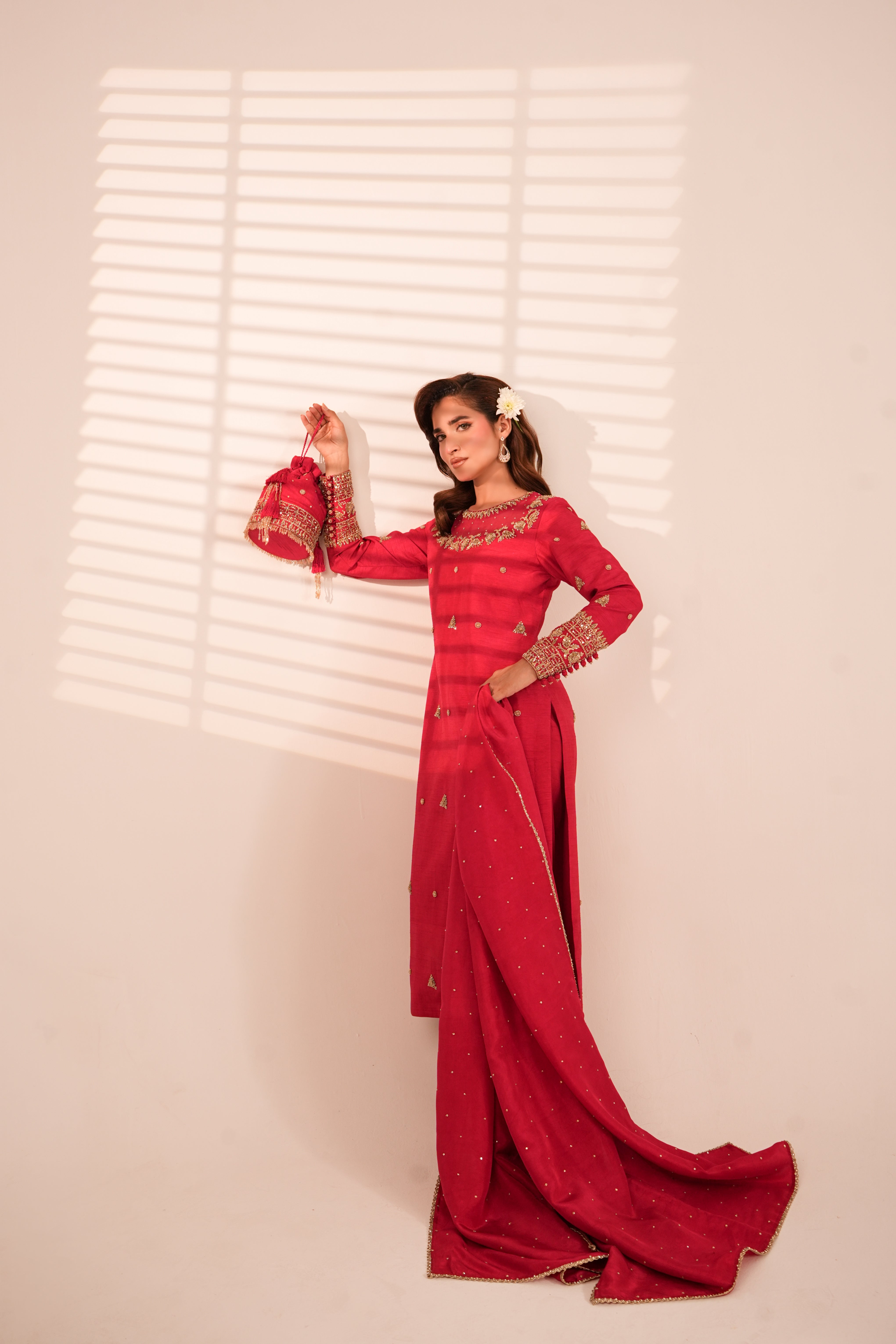 A woman models a long red traditional dress with gold embroidery and beadwork, standing against a light beige wall with window blind shadows.