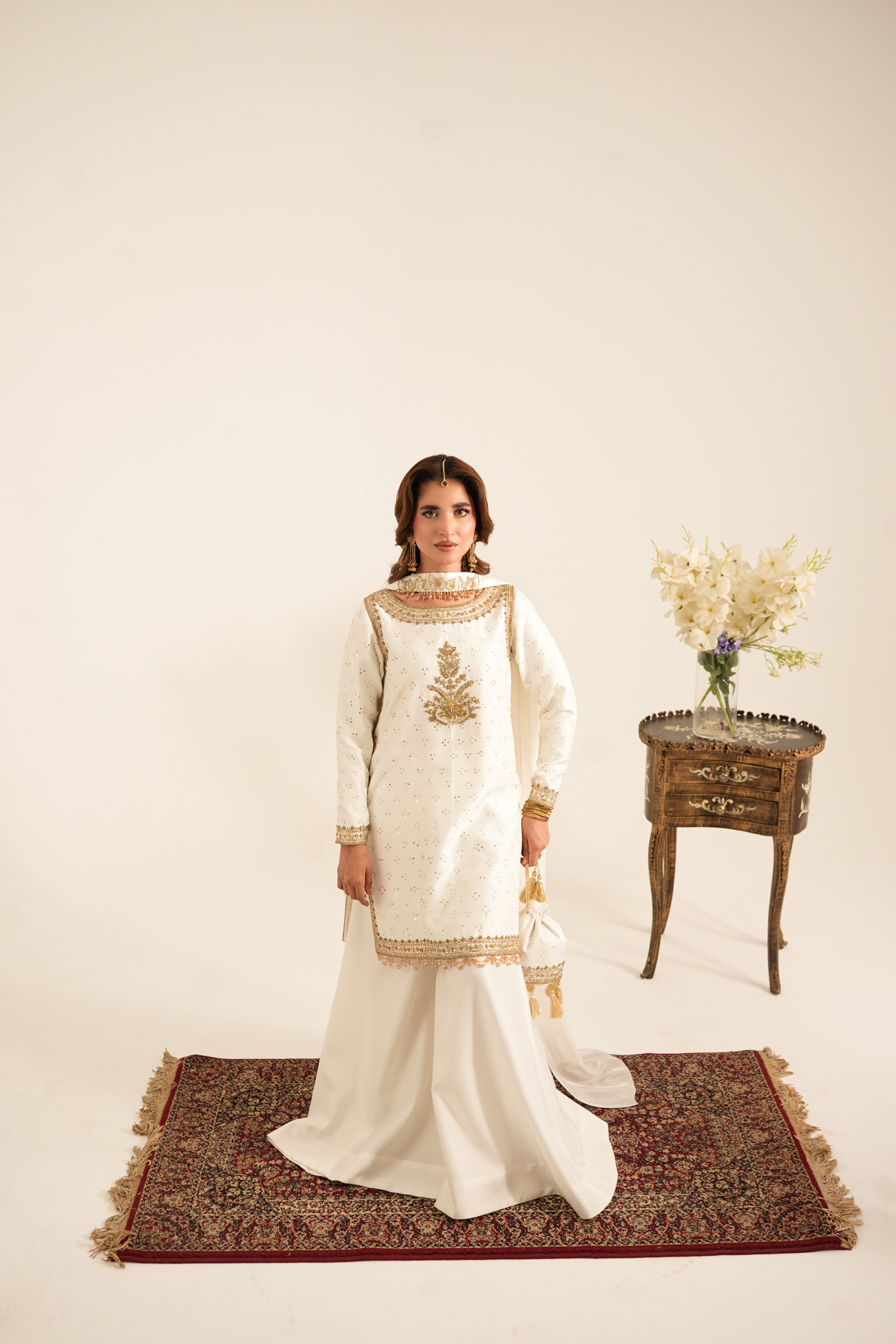 A woman stands on a patterned rug wearing a traditional white embroidered kurta set with a long skirt. She is posed in a studio setting with a small wooden side table holding a vase of flowers in the background.