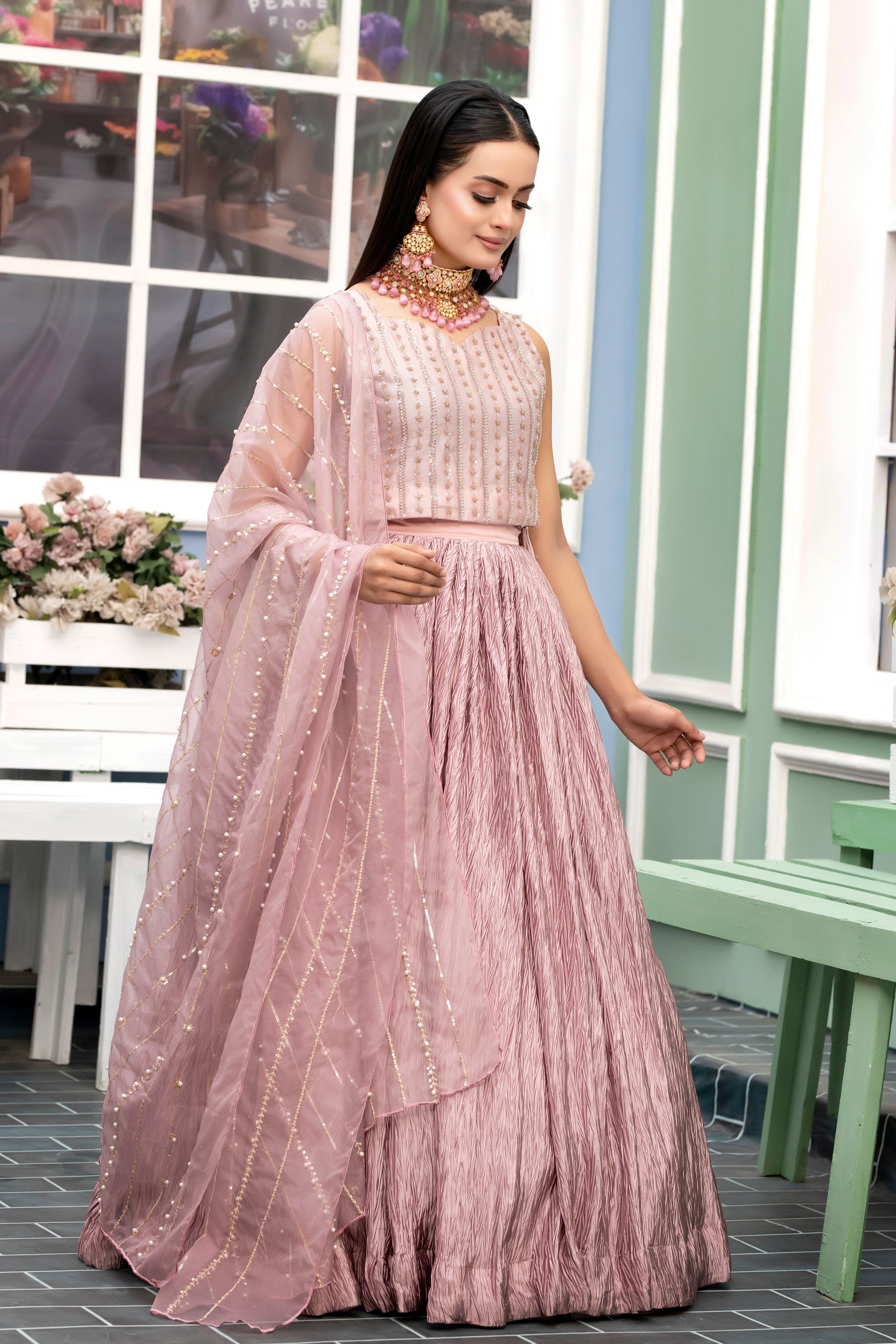 Woman in a pink traditional outfit standing in a decorative outdoor setting with flowers and a bench.