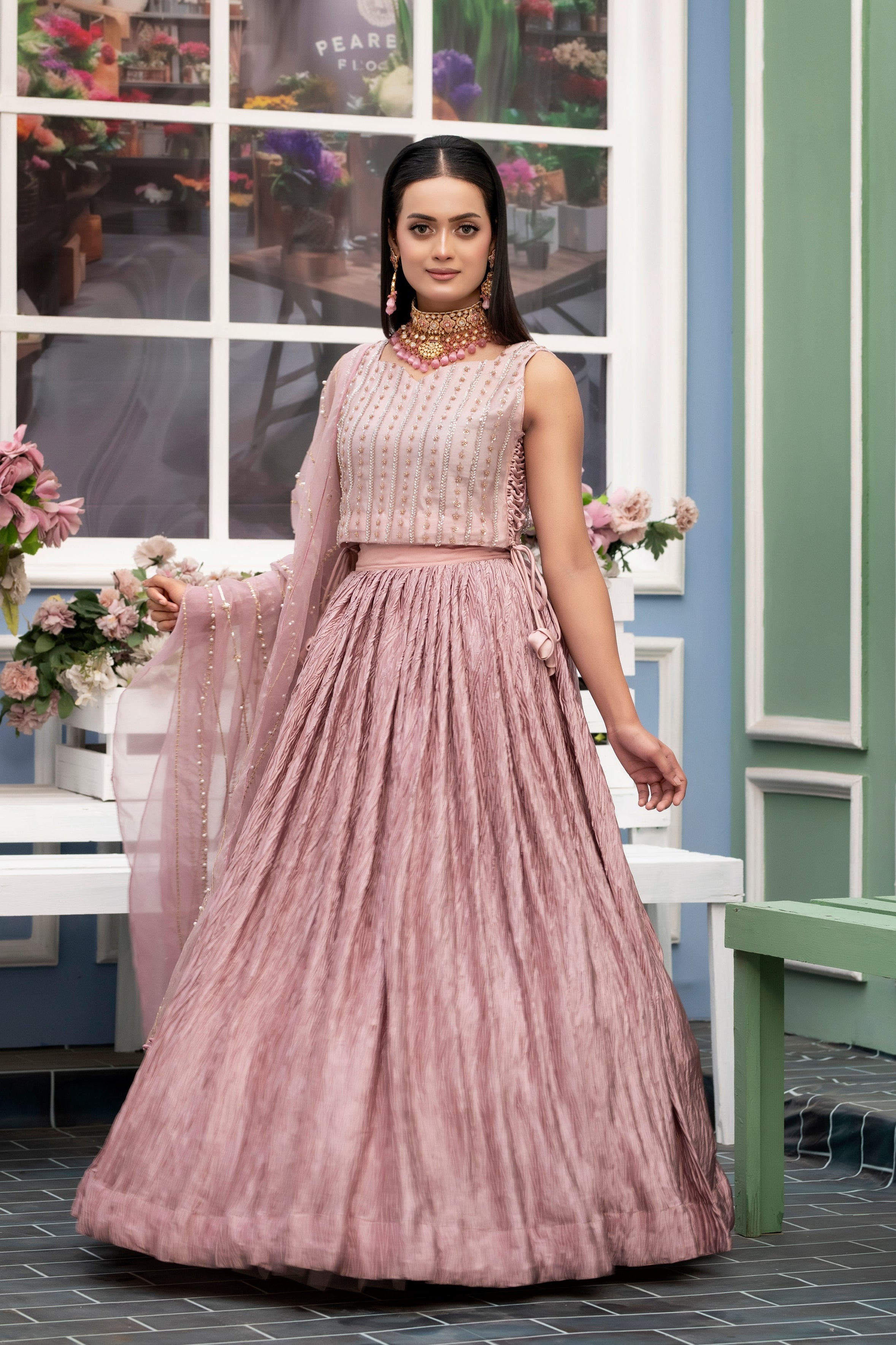 Woman in a pink traditional outfit standing in a decorated room with flowers and furniture.