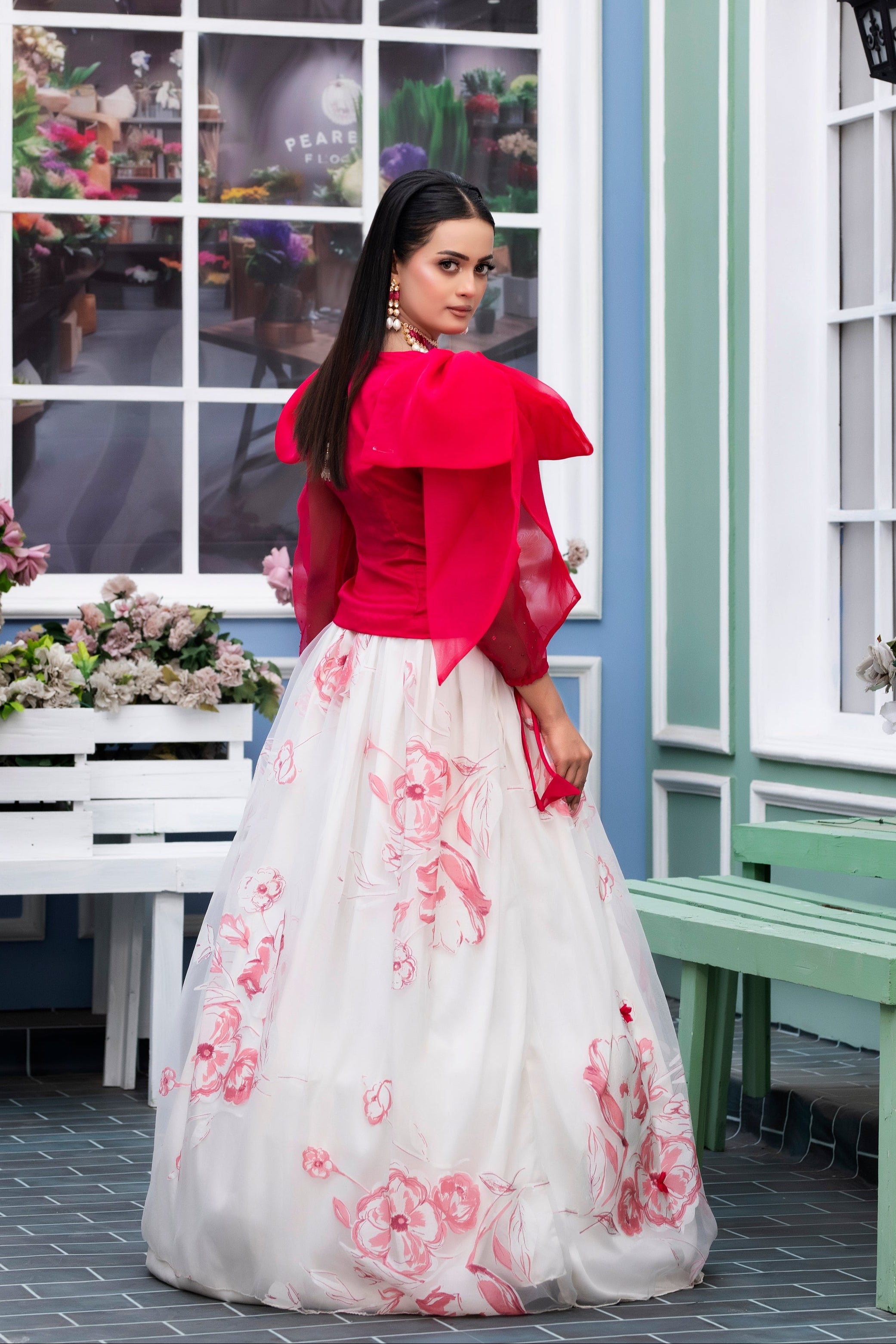 Woman in a red top and white floral skirt standing in a decorative setting with flowers and benches.