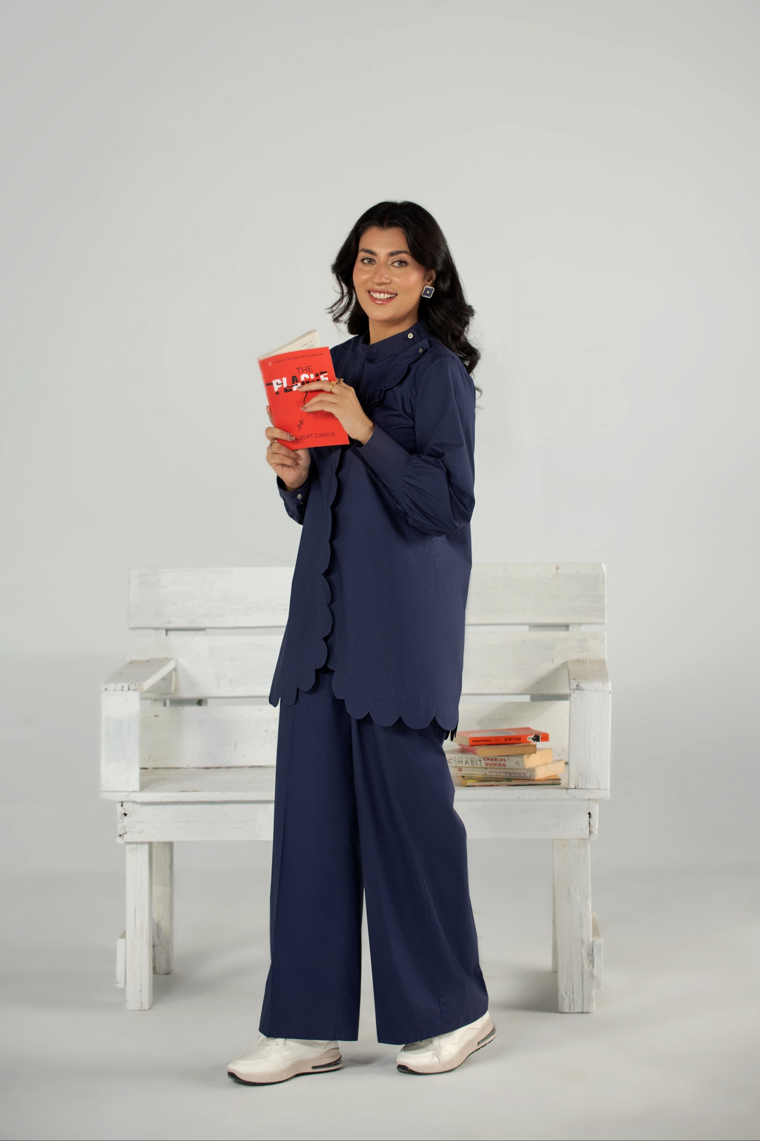 Woman in navy outfit holding a red book on a white bench against a gray background