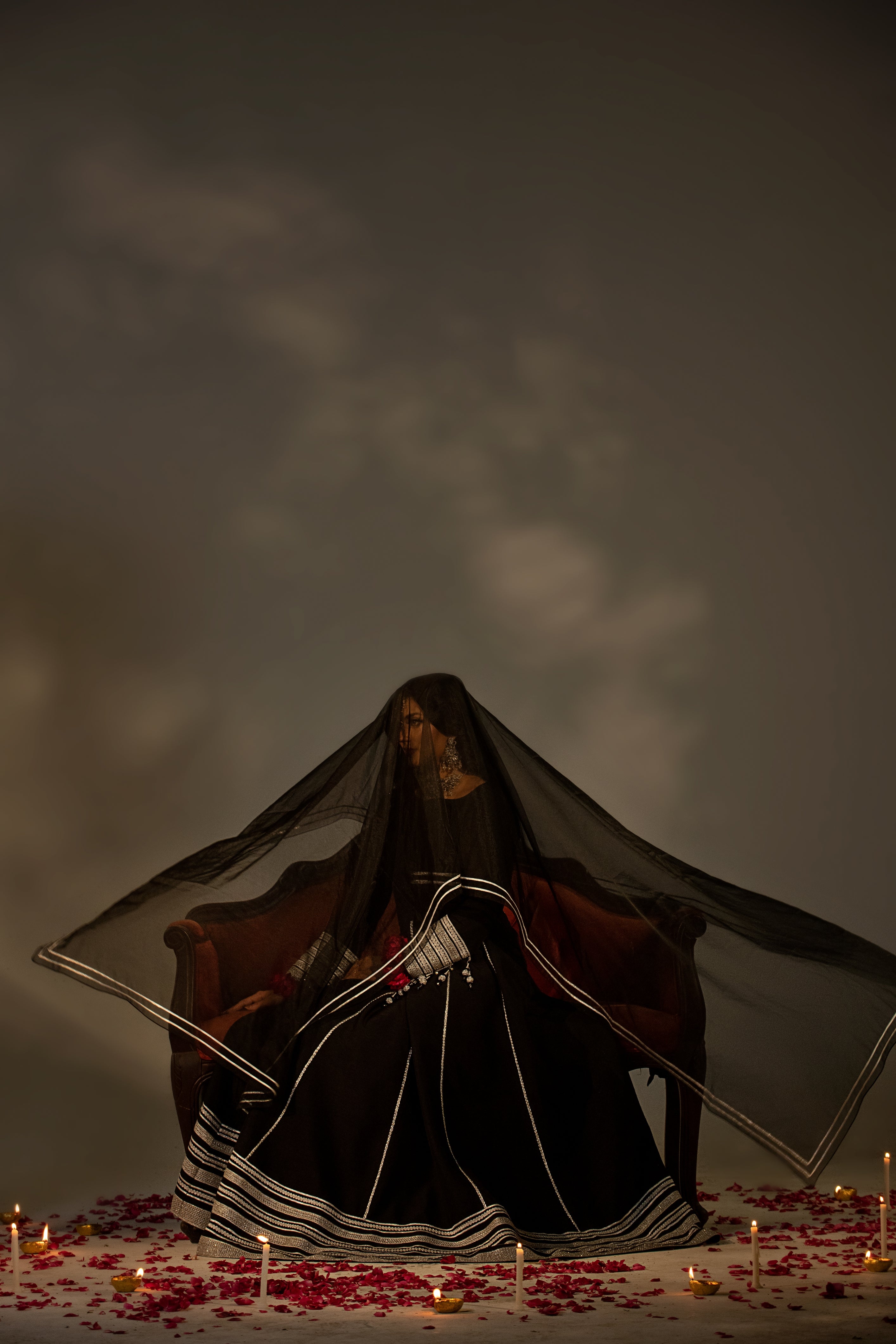 Woman in a dark dress with a sheer overlay sitting on a bench with candles and rose petals on a dark background
