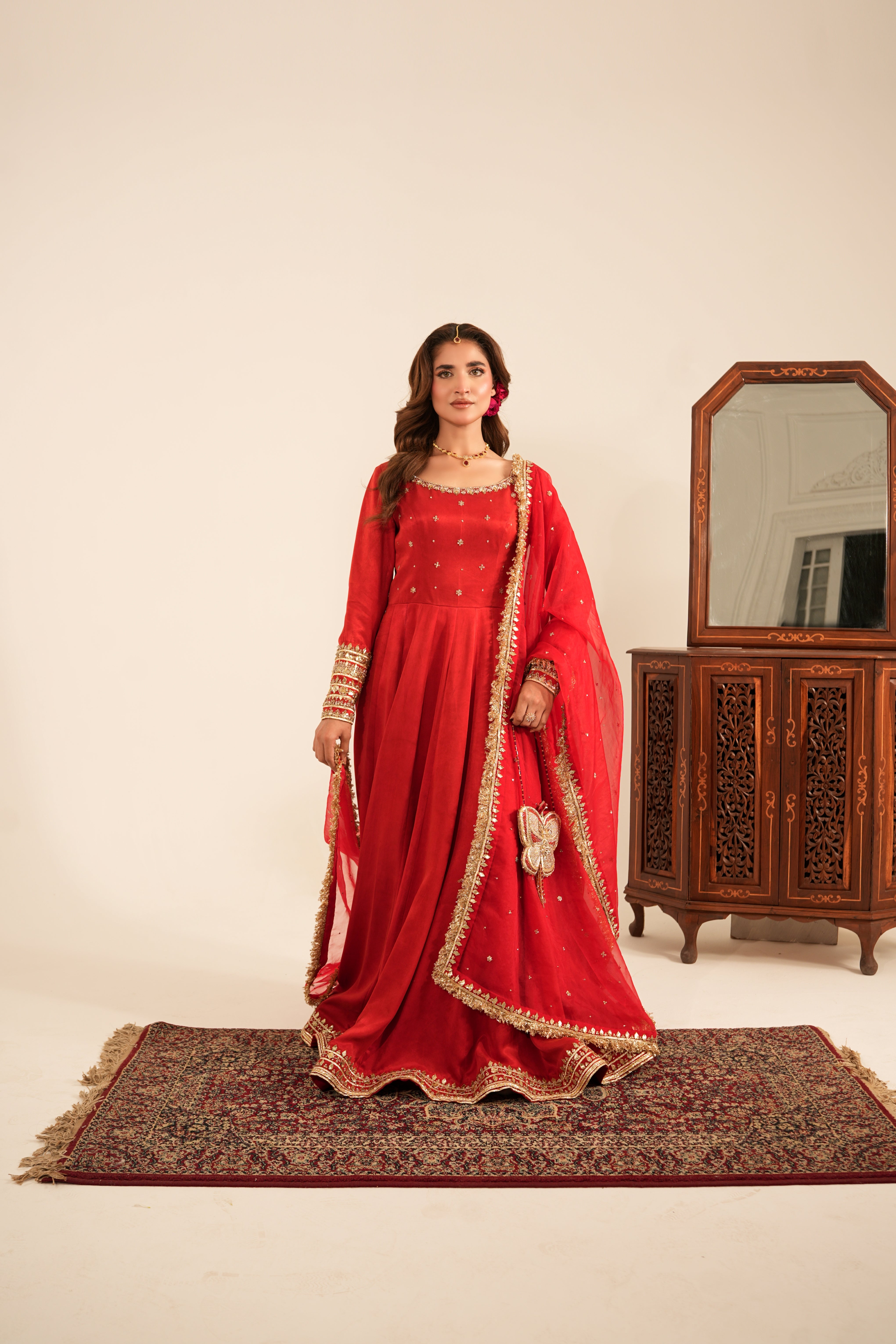A woman models a traditional red Indian salwar kameez suit with gold embroidery, standing on a patterned rug in front of a wooden cabinet with a mirror.