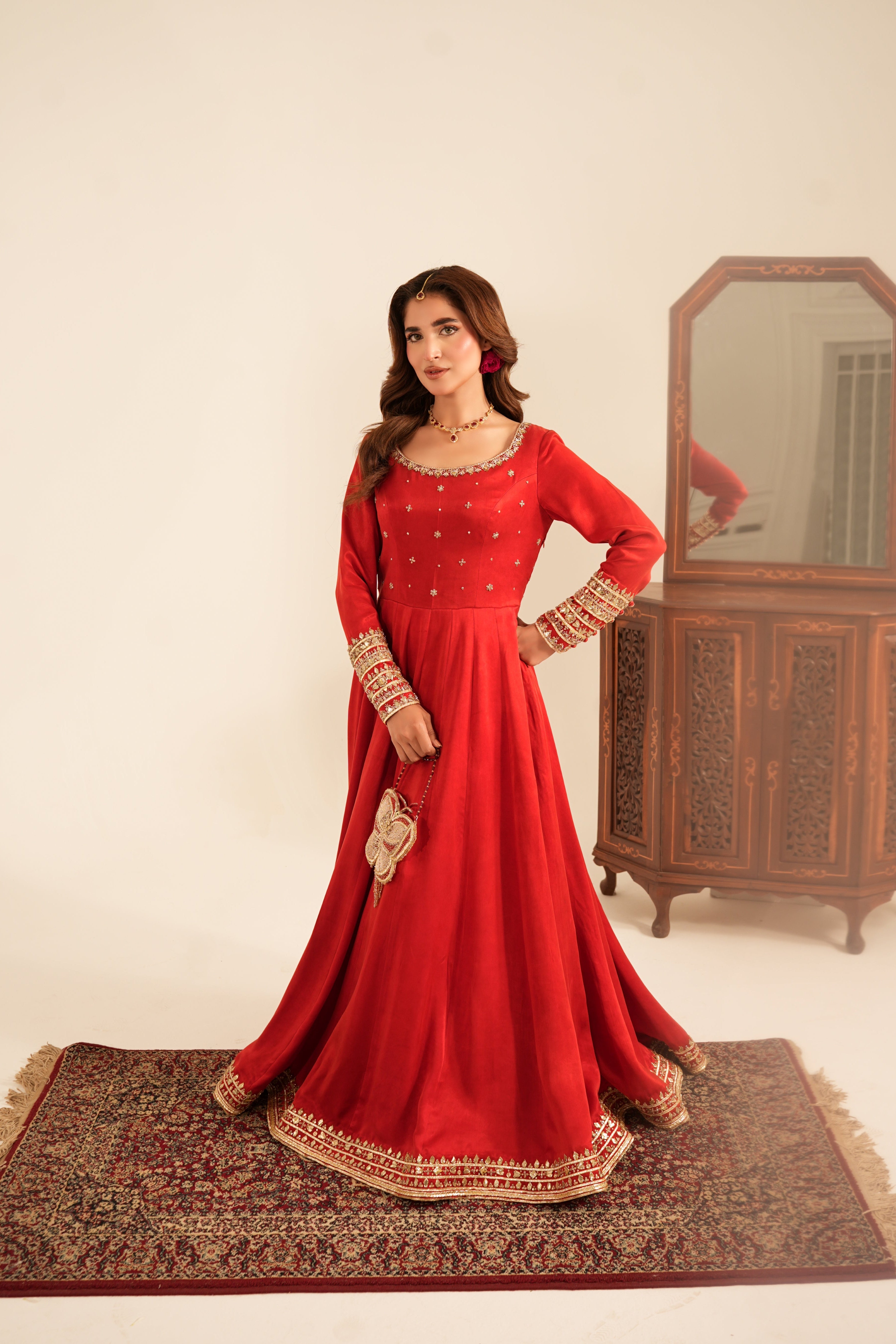 A woman models a traditional red long gown with gold embroidery, standing on a patterned rug in front of a wooden cabinet and mirror.