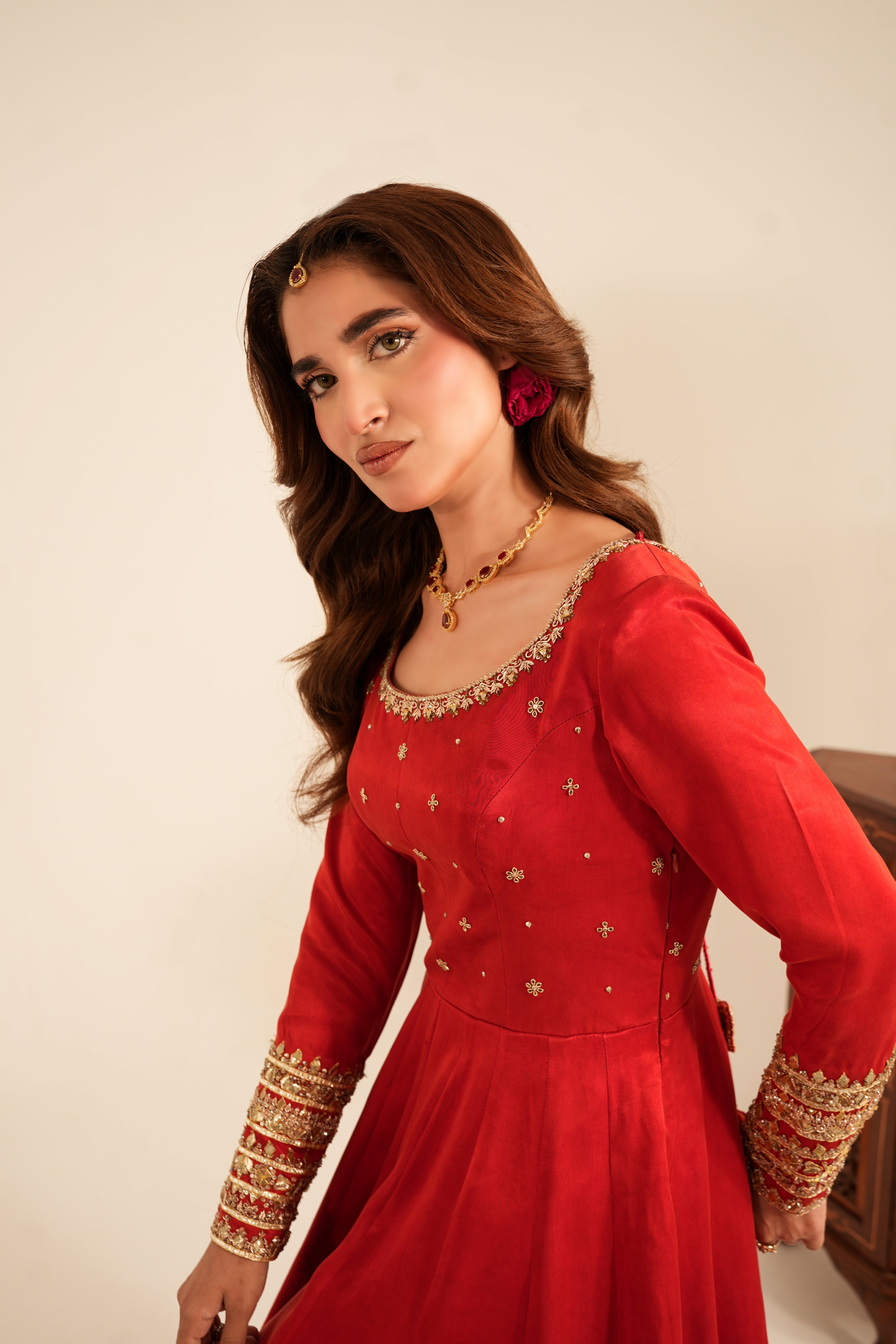 A woman wearing a red long-sleeve Anarkali-style dress with gold embroidery and embellishments, posed against a plain beige wall.