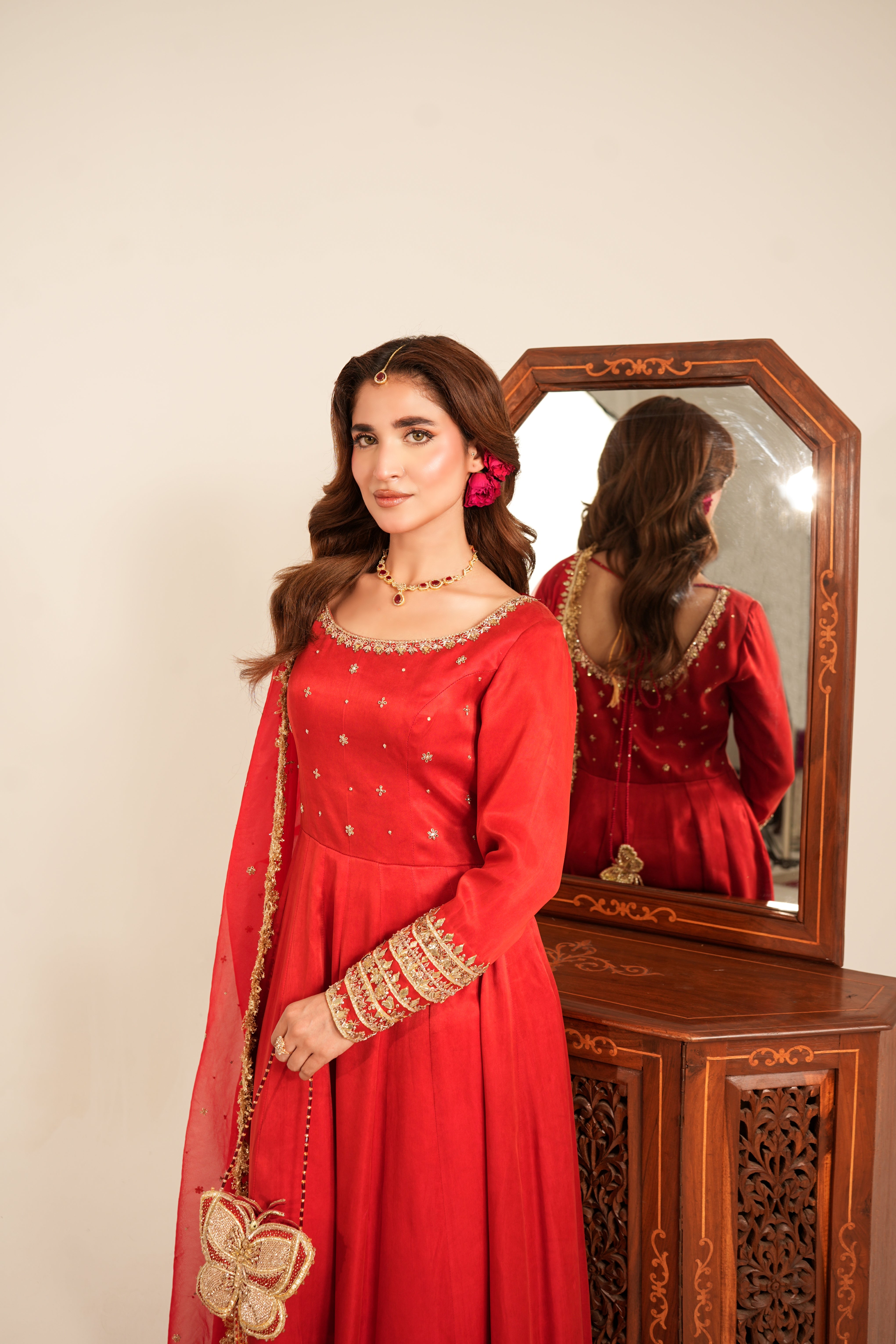 A woman wearing a bright red traditional embroidered dress stands beside a wooden mirror and carved cabinet, with her reflection visible in the mirror.