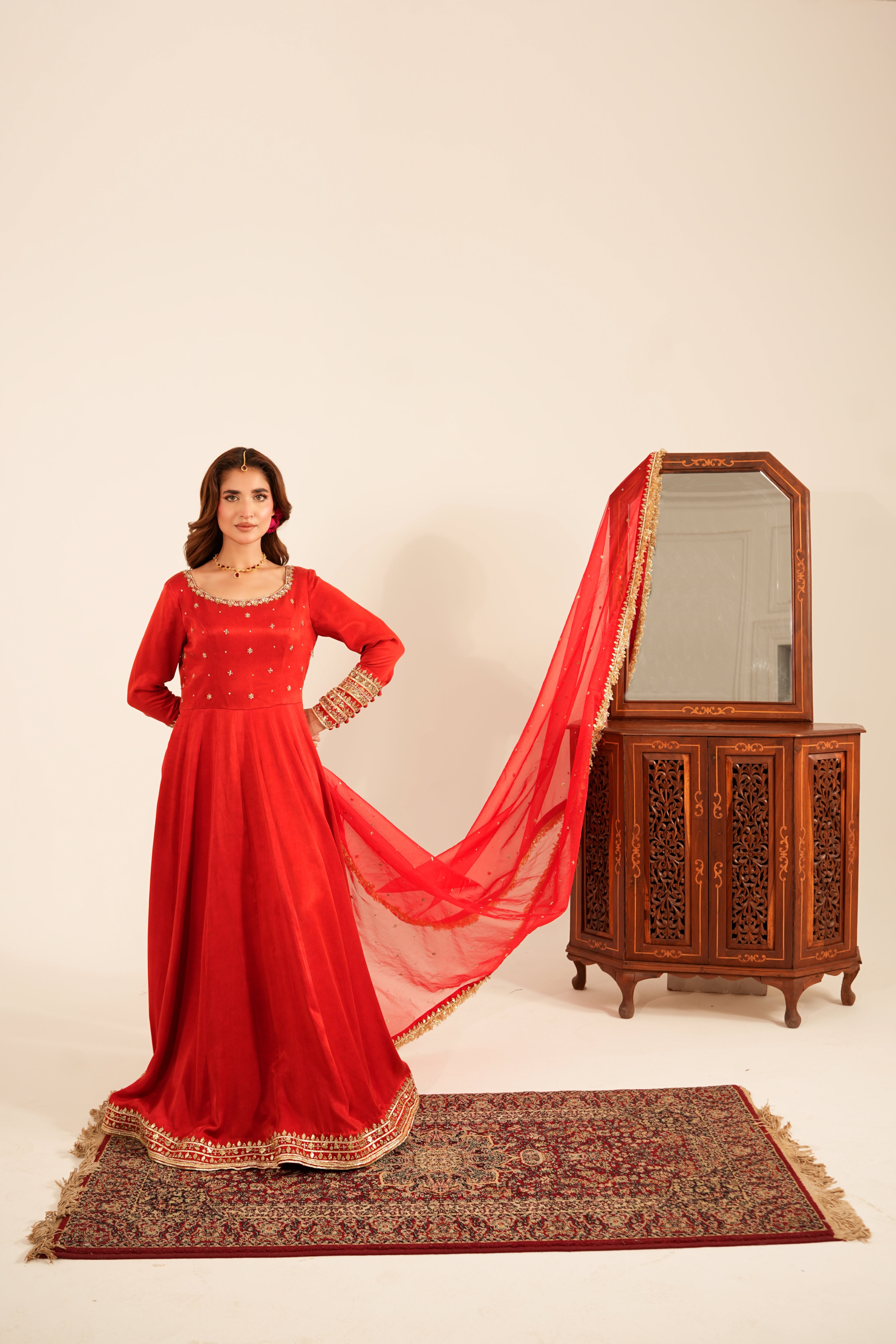 A woman models a traditional red Indian salwar kameez outfit with a matching dupatta, standing on a patterned rug next to a carved wooden cabinet with a mirror, against a plain beige background.
