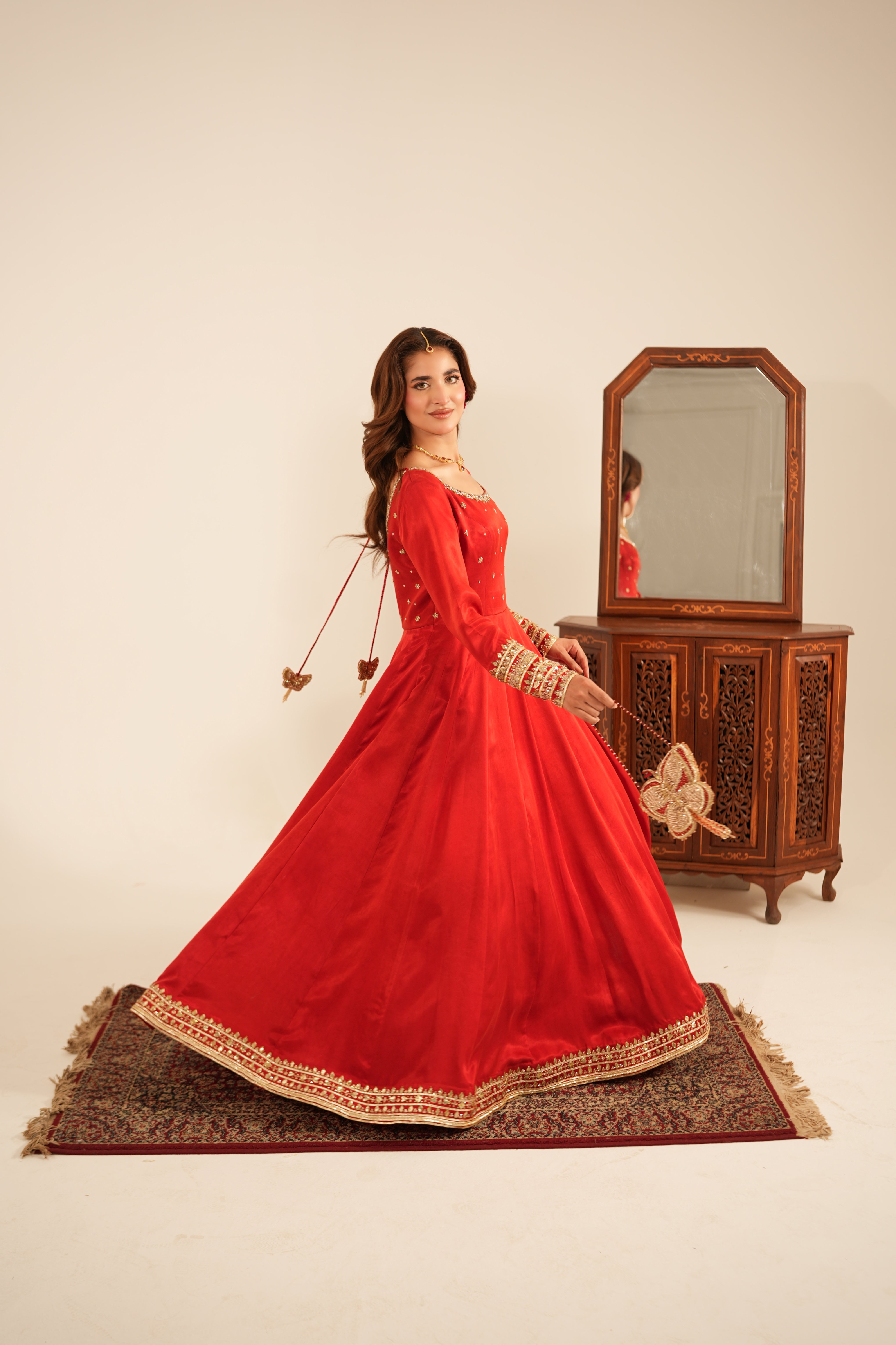 A woman wearing a vibrant red traditional long gown with gold embroidery stands on a patterned rug in front of a wooden cabinet and mirror.
