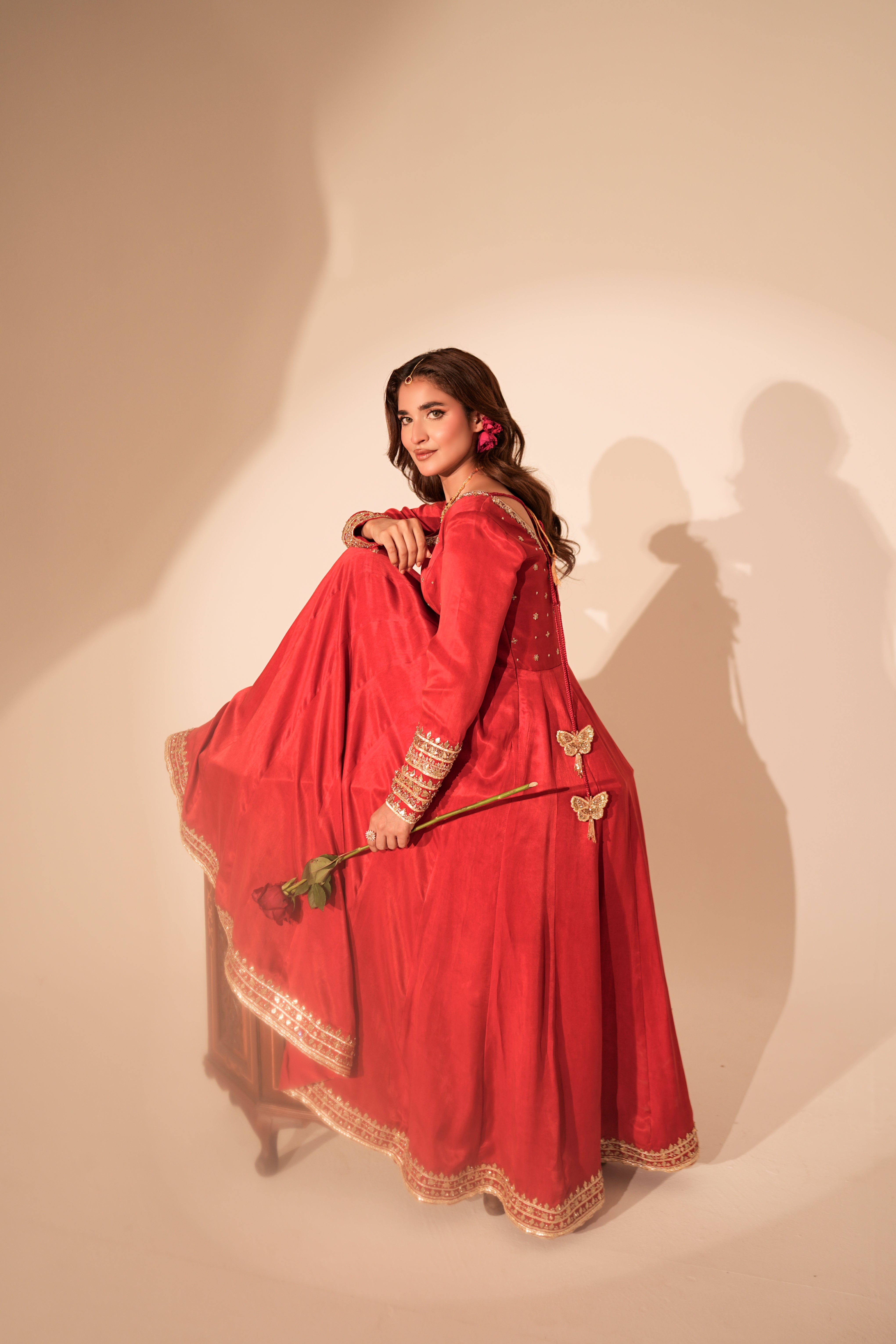 A woman wearing a vibrant red traditional dress with gold embroidery and a flared skirt, holding a green stem against a warm beige studio backdrop with dramatic shadows.