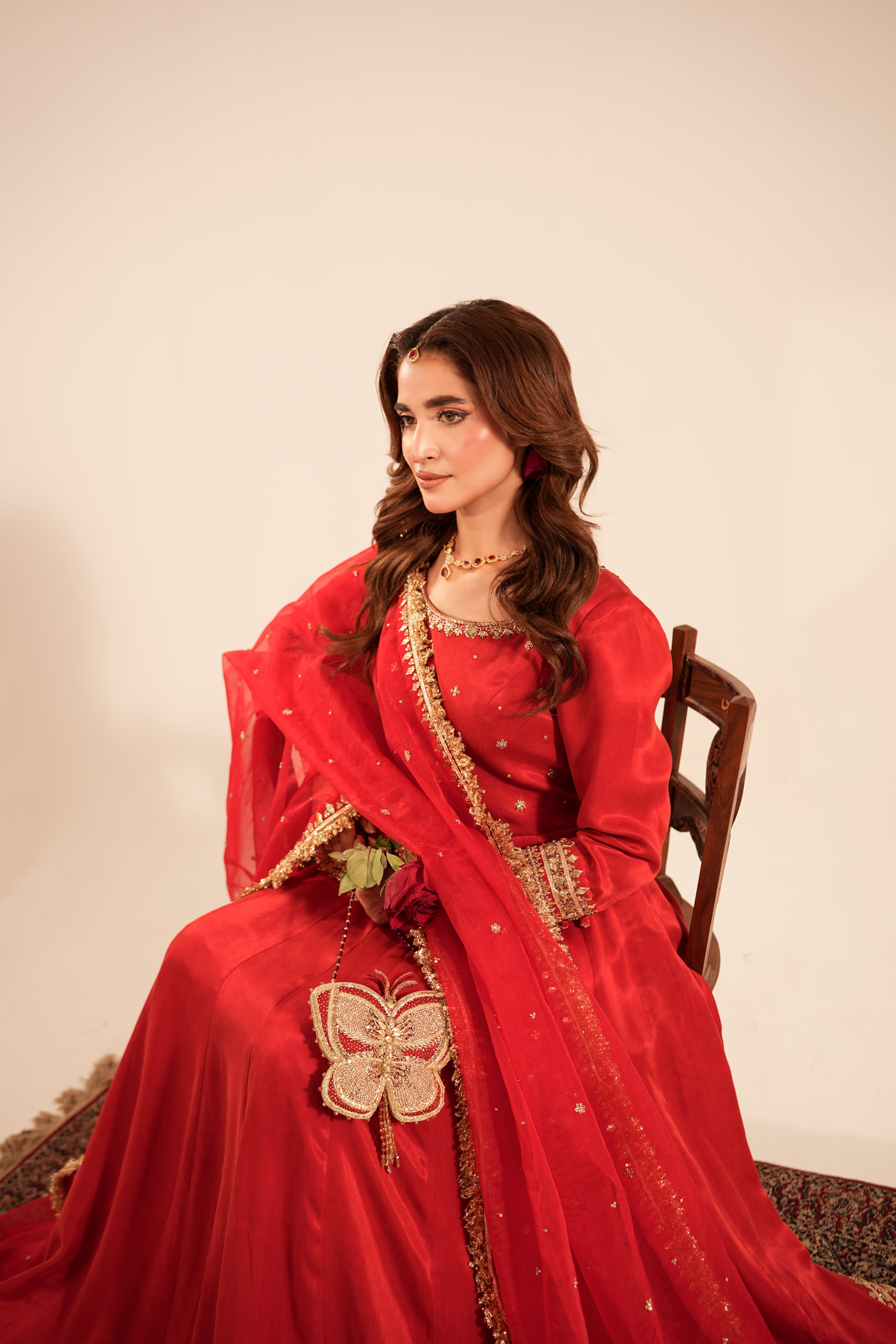 A woman seated on a wooden chair wearing a flowing red traditional outfit with gold embroidery and a sheer dupatta, photographed against a plain light beige wall.