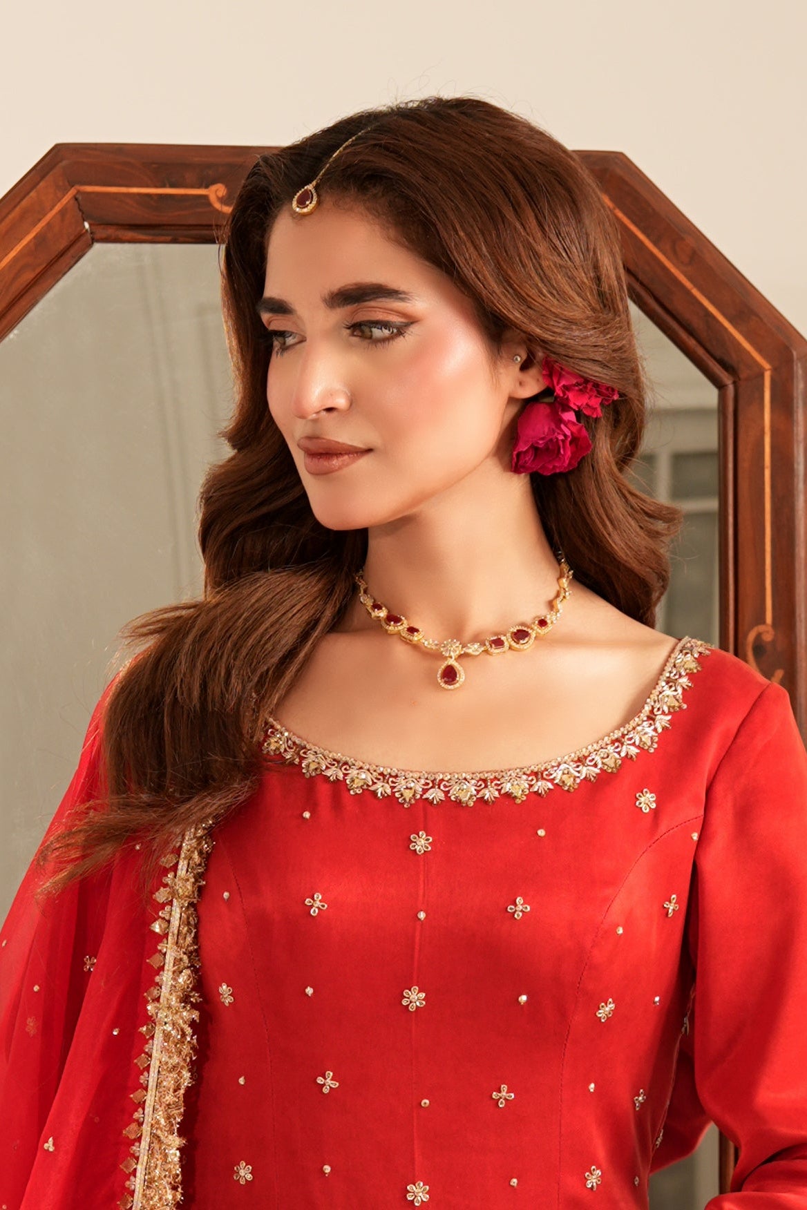 A woman wearing a red traditional outfit with gold embroidery and a matching dupatta, standing in front of a wooden-framed mirror against a plain beige wall.