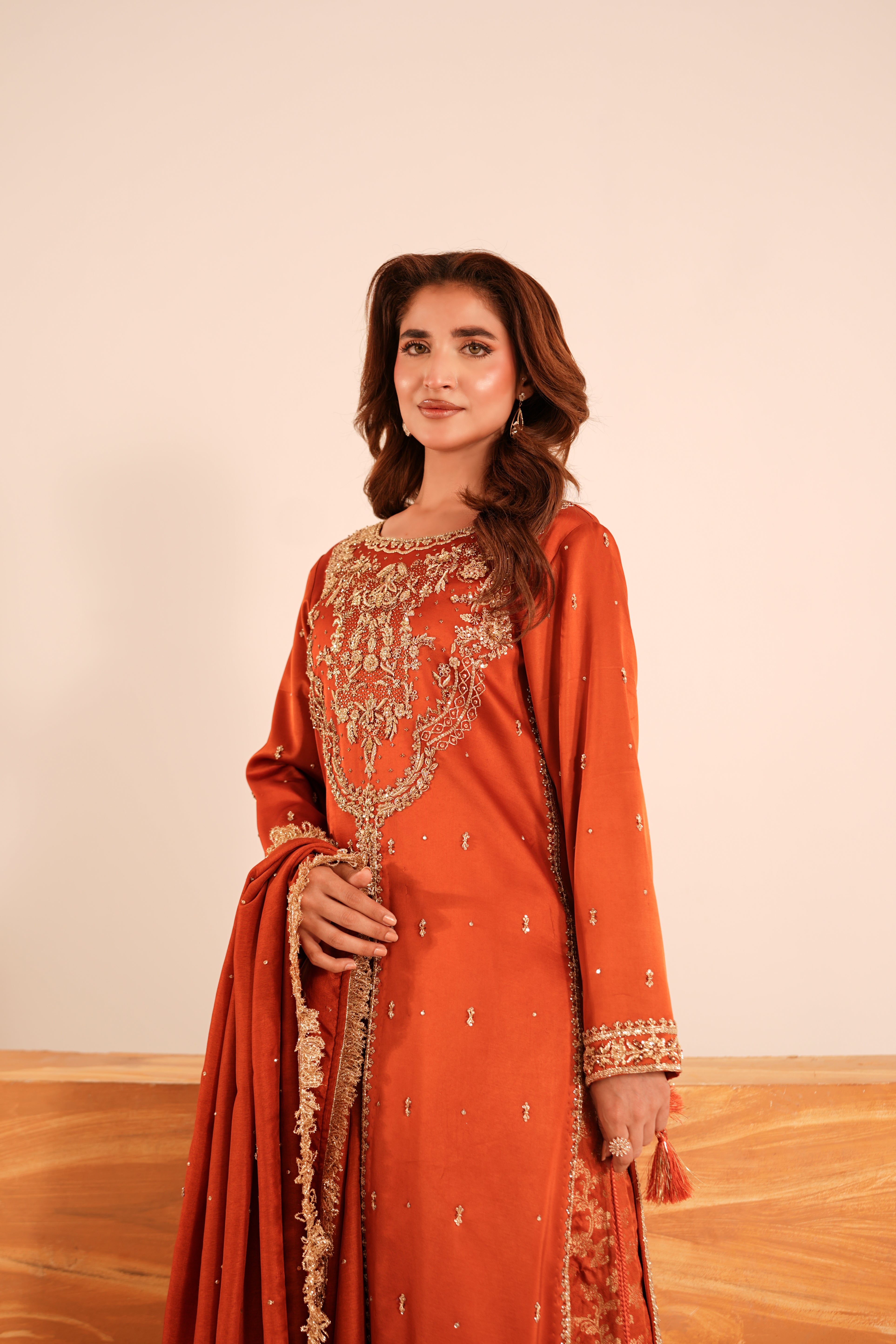 A woman modeling a traditional rust-orange salwar kameez suit with intricate gold embroidery and a matching dupatta, set against a warm beige wall and light wood floor.