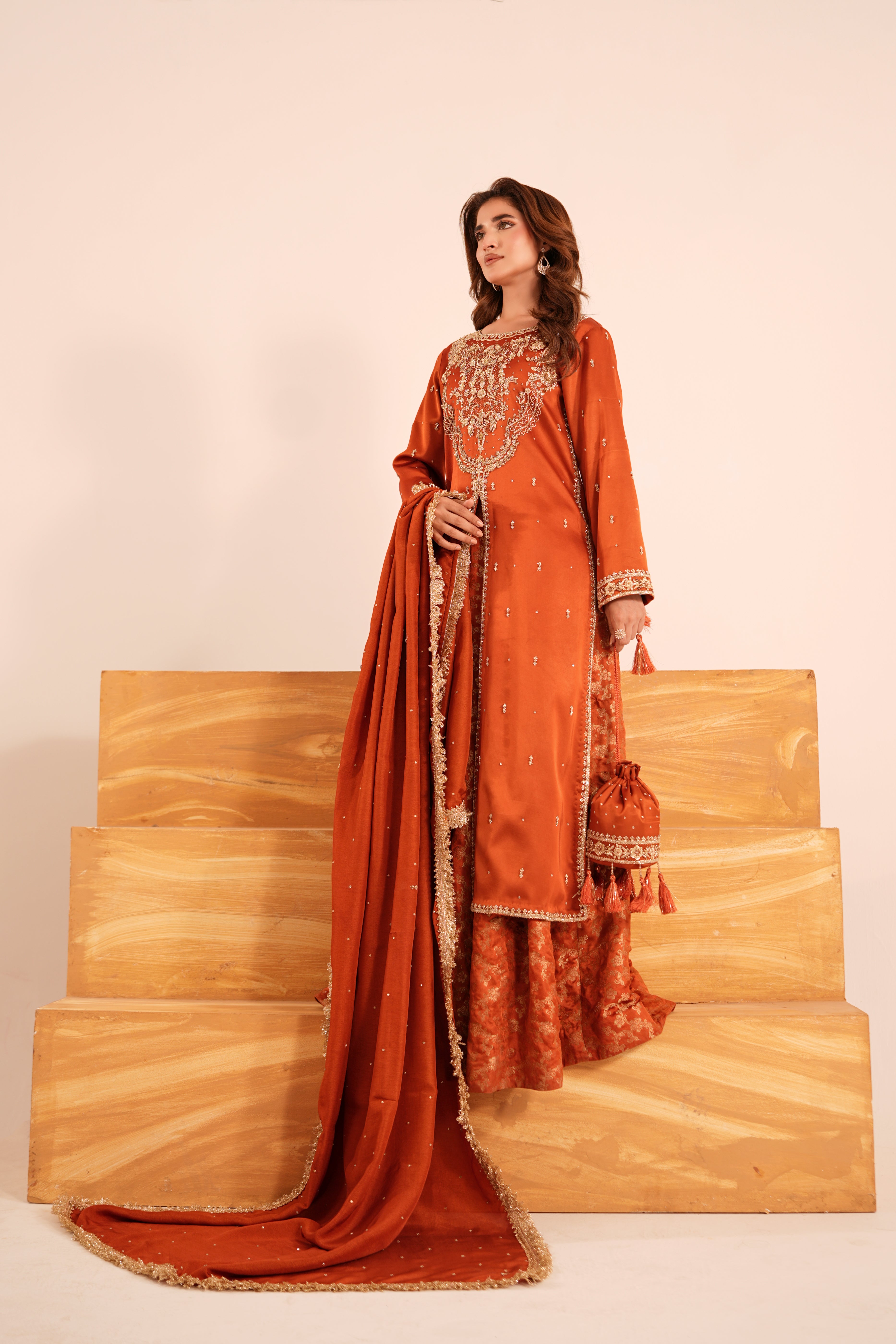 A woman models an orange traditional South Asian outfit with intricate gold embroidery and a matching dupatta, standing on wooden steps against a plain beige background.