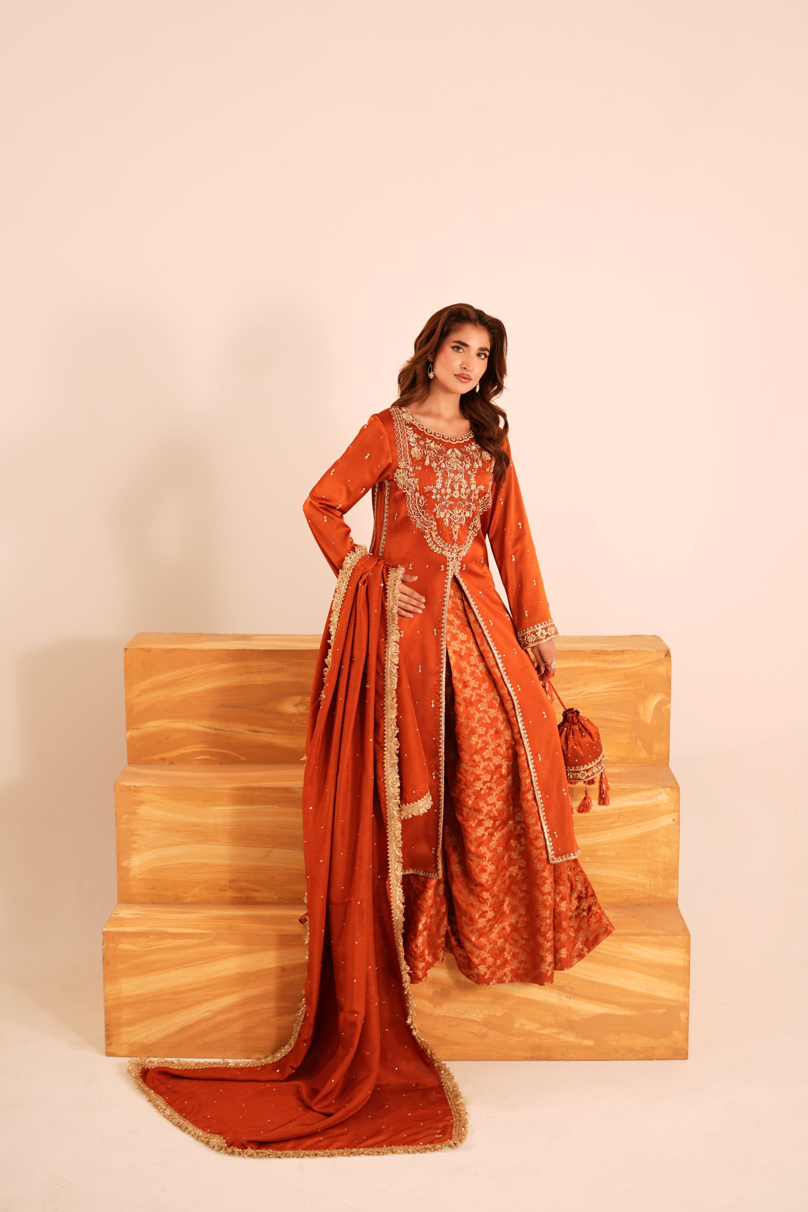 A woman models an orange traditional Indian outfit with intricate gold embroidery and a matching dupatta, standing on wooden steps against a beige background.