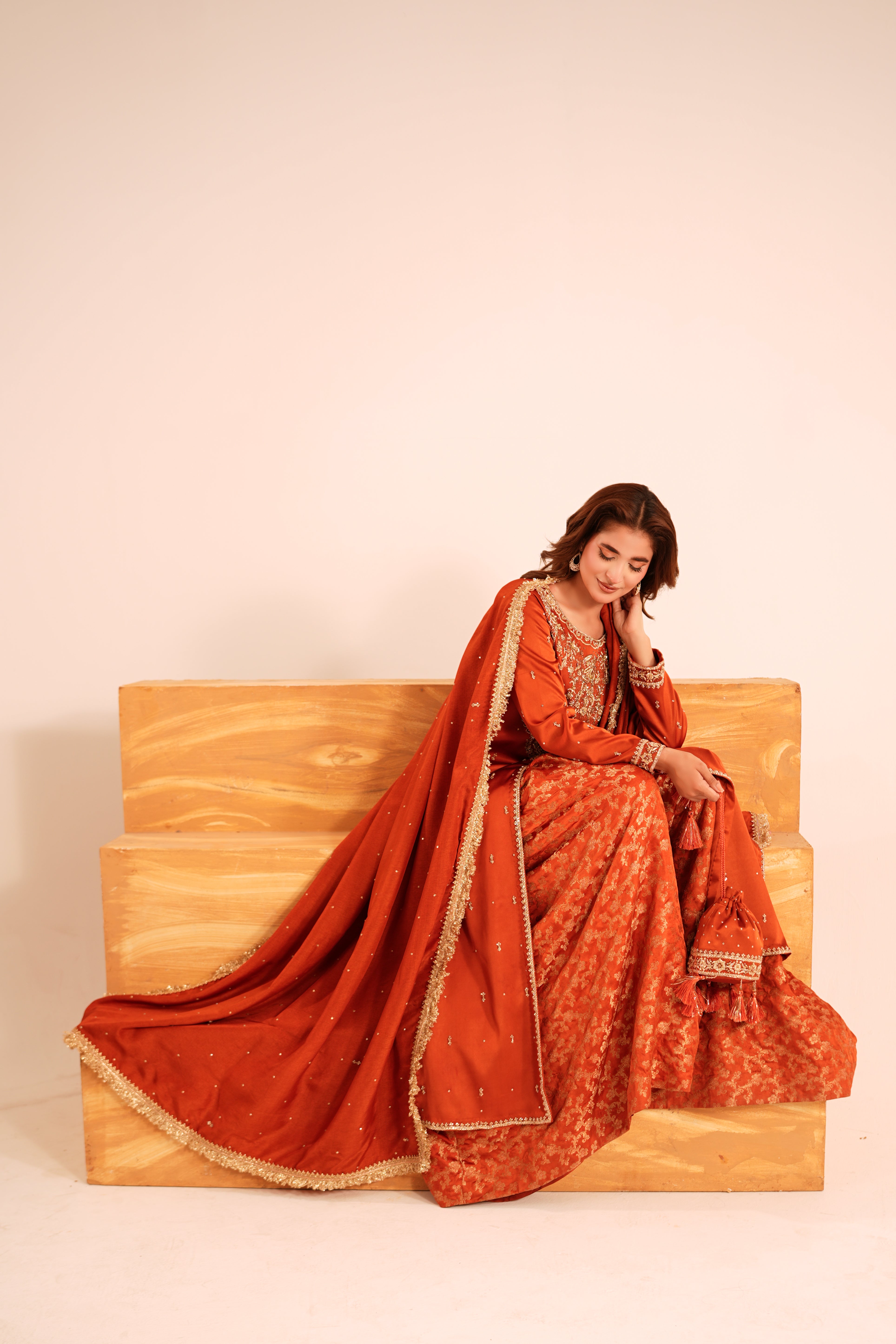 A woman wearing a traditional Indian salwar kameez outfit in rust orange and gold, sitting on a wooden bench against a plain beige background.