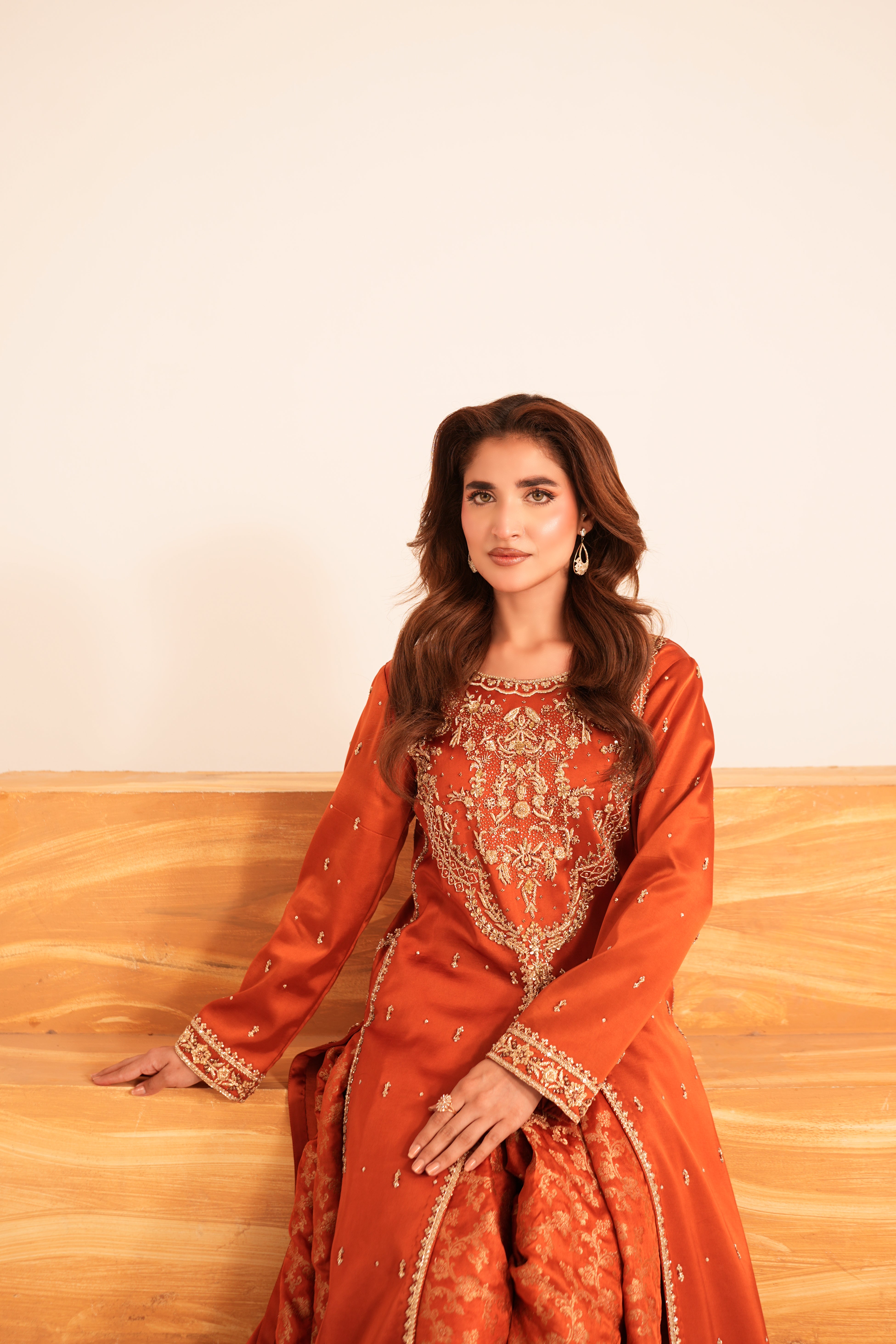 A woman wearing a rust-orange embroidered traditional outfit with gold detailing, seated on a light wood floor against a plain off-white wall.