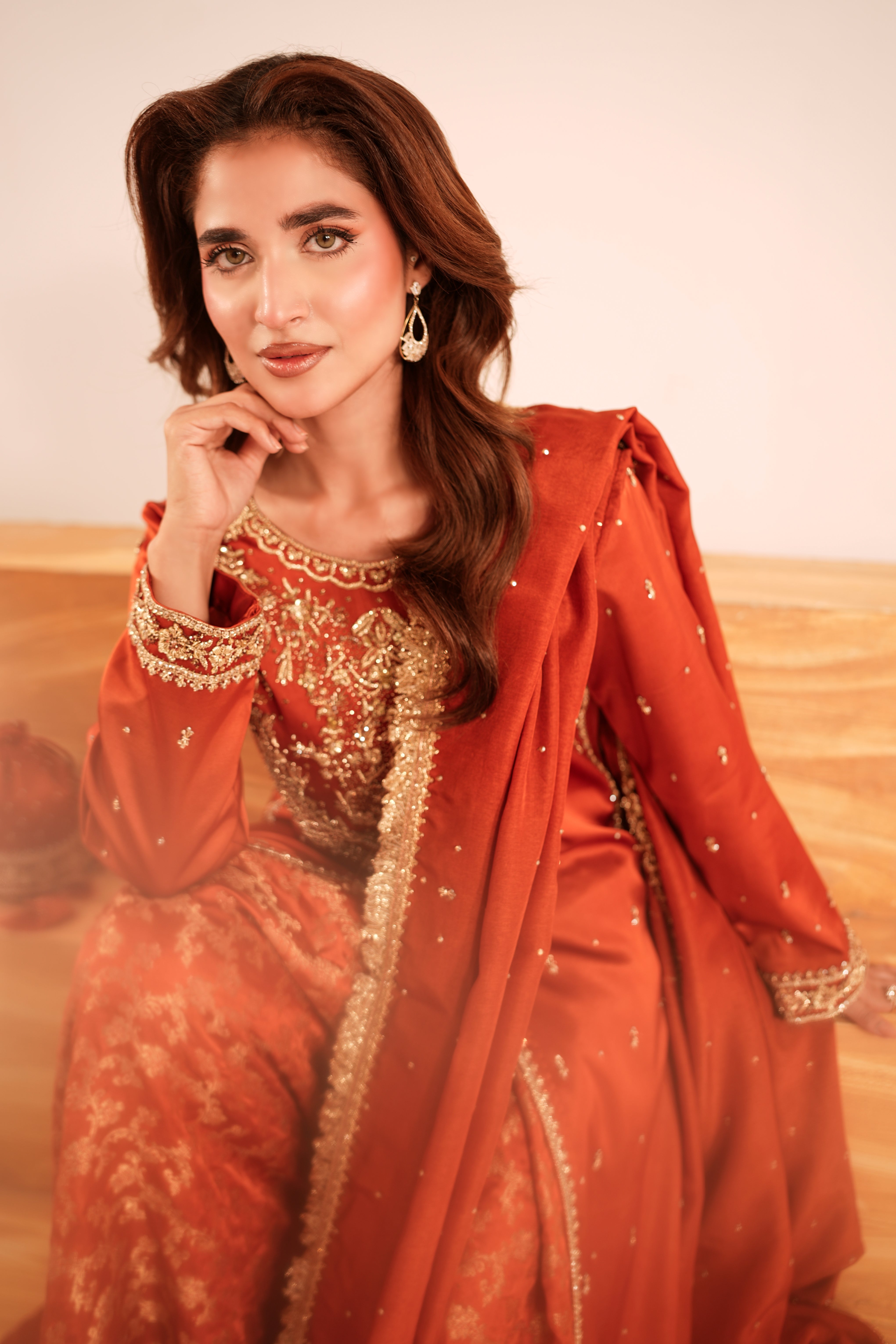 A woman wearing a traditional South Asian outfit in rust orange and gold, posing indoors against a neutral beige background.
