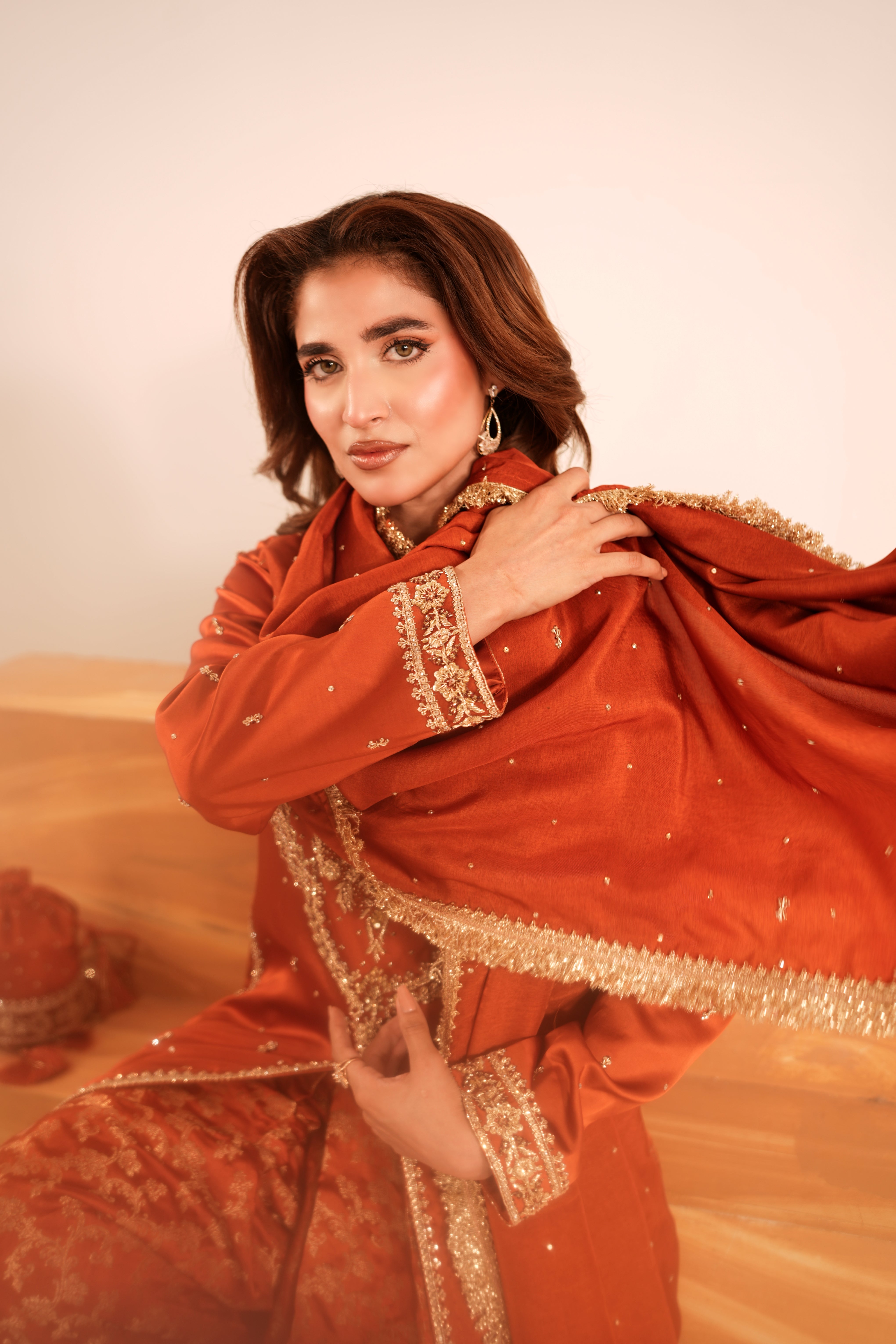 A woman wearing a traditional rust-orange embroidered outfit with a matching dupatta draped over her shoulders, posed against a warm beige background.