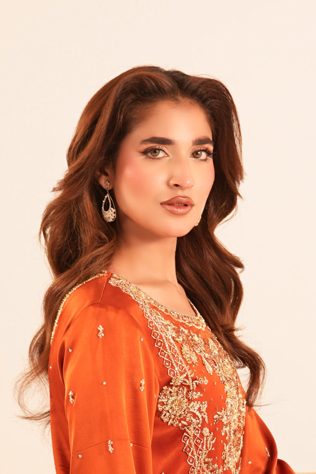 A woman with long wavy brown hair is shown from the waist up, wearing a bright orange traditional outfit with gold embroidery, set against a plain cream-colored background.